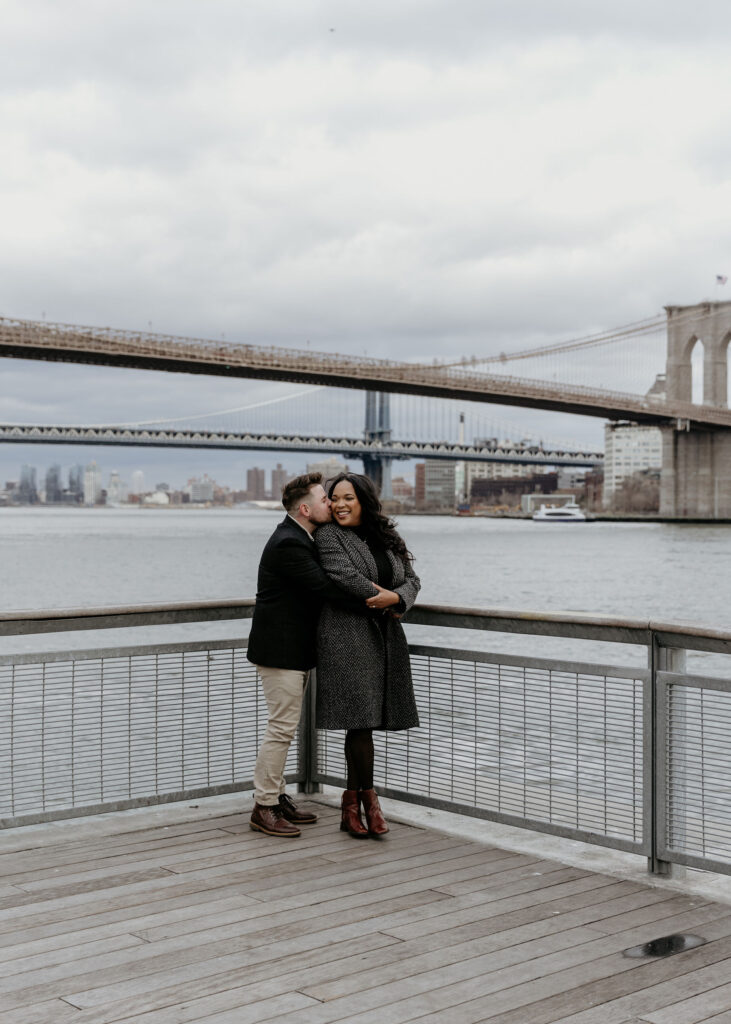 New york city couple hugging and snuggling in south seaport street, pier 17 in downtown manhattan with the brooklyn bridge in the background